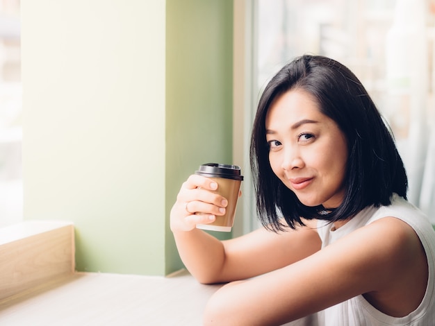 La mujer asiática bebe el café caliente en el café con la luz caliente.