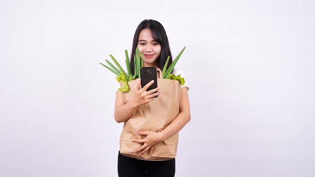 Mujer asiática con bolsa de papel de verduras frescas y sosteniendo un teléfono con superficie blanca aislada