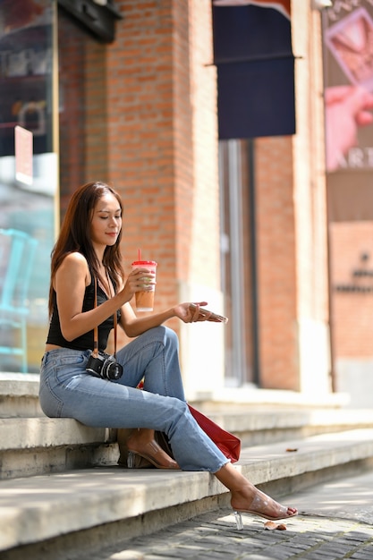 Mujer asiática con bolsas de compras, cámara retro, café para llevar y teléfono inteligente en mano sentado en las escaleras