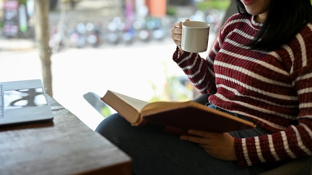 Mujer asiática brillante leyendo un libro mientras se relaja tomando café en la imagen recortada del café