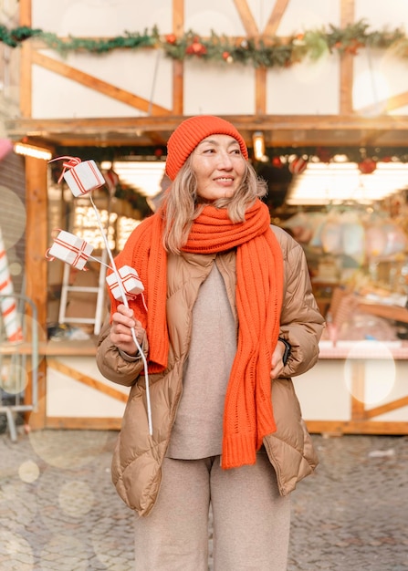 Mujer asiática con bufanda naranja y sombrero caminando en el mercado navideño decorado con luces navideñas