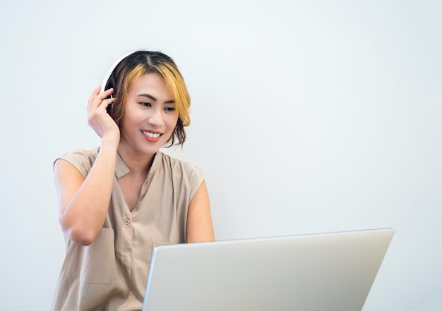 Mujer asiática con cabello corto de moda usando auriculares blancos mientras trabaja con una computadora portátil independiente mirando la pantalla sobre fondo de pared blanca con espacio de copia Concepto de reunión en línea