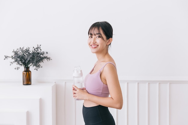 Mujer asiática con cabello largo y negro atado feliz sonriendo a la cámara usando ropa deportiva y sosteniendo una botella de agua de pie sobre un fondo blanco Imagen de estilo de vida activo con espacio de copia