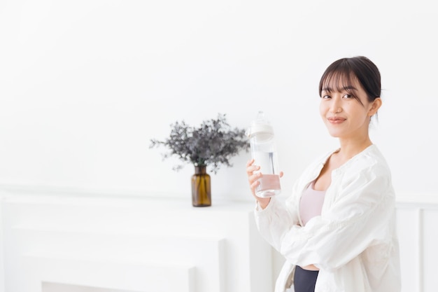 Mujer asiática con cabello largo y negro atado usando ropa deportiva y una chaqueta deportiva blanca Feliz sonriendo a la cámara y sosteniendo una botella de agua de pie sobre un fondo blanco Imagen con espacio de copia