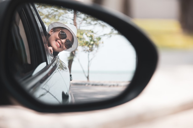 La mujer asiática en la camisa blanca está mirando en el espejo de la vista lateral y está sonriendo mientras que se sienta en su coche, concepto del viaje.