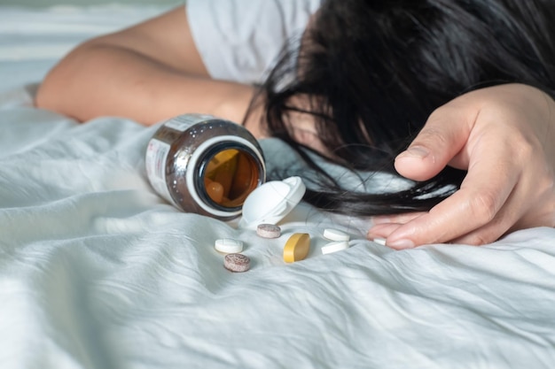 Una mujer asiática con camisa blanca yace inconsciente en una cama blanca después de una sobredosis de píldoras para bajar de peso, seleccione el enfoque en las píldoras para contar una historia