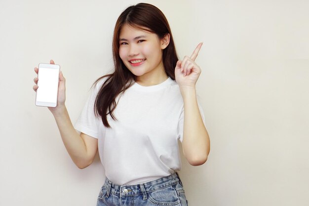 La mujer asiática con camiseta blanca sonríe mientras sostiene el teléfono móvil y señala hacia arriba