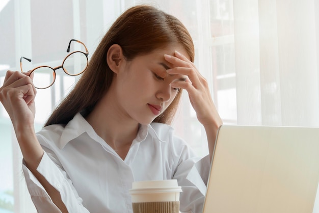 Mujer asiática cansada o deprimida que se sienta detrás de su computadora portátil con sus manos que sostienen las lentes