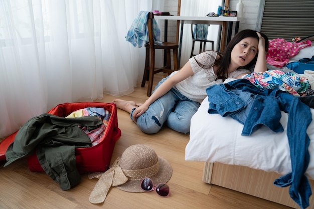 Mujer asiática cansada sentada en el suelo apoyada en una cama blanca con ropa de maleta de viaje dentro. una joven que se siente aburrida mientras empaca el equipaje es difícil pensar qué ponerse para las vacaciones de verano.