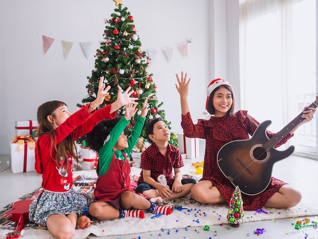 La mujer asiática celebra la Navidad al acercar la guitarra al niño. Los niños juegan juntos el día de Navidad con el árbol de navidad.