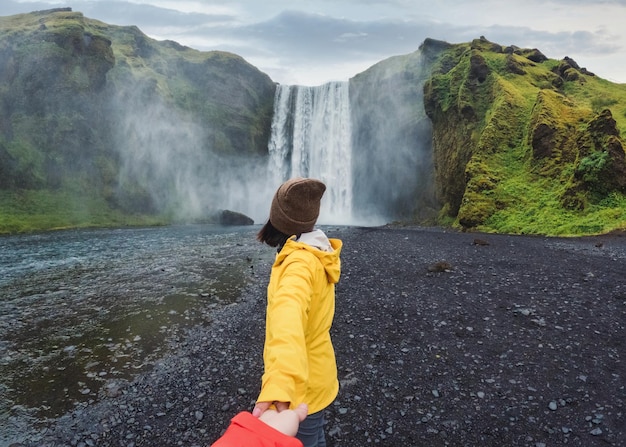 Mujer asiática cogida de la mano con una pareja frente a la cascada de Skogafoss que fluye sobre un acantilado en verano en Islandia