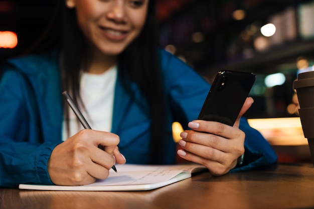 Mujer asiática complacida recortada haciendo notas en el planificador y usando el teléfono celular mientras está sentado en la cafetería