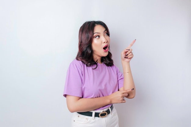 Una mujer asiática conmocionada con una camiseta morada lila apuntando al espacio de la copia a su lado aislada por el fondo blanco