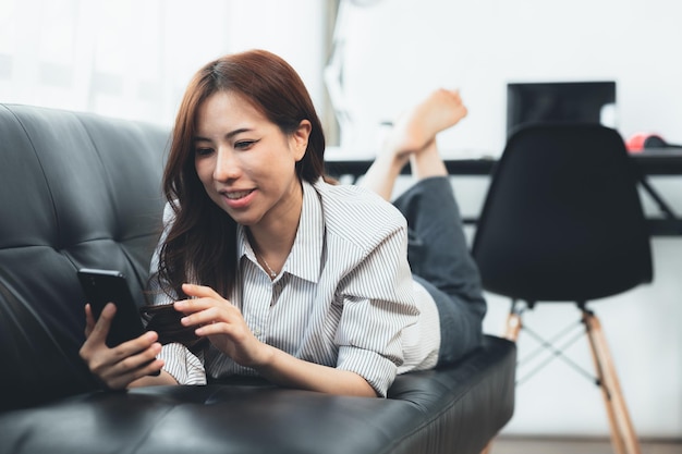 Mujer asiática descansando en el sofá que está enviando mensajes de texto a través de la aplicación en un teléfono inteligente con un amigo que está tomando un descanso de fin de semana después de un duro día de trabajo Ideas de relajación de vacaciones