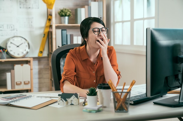 mujer asiática diseñadora de interiores bostezando mano cubierta boca sentada en un estudio moderno y luminoso. elegante joven ingeniera mirando fijamente a la computadora sintiéndose cansada exhausta con exceso de trabajo soñolienta sentada en la oficina.