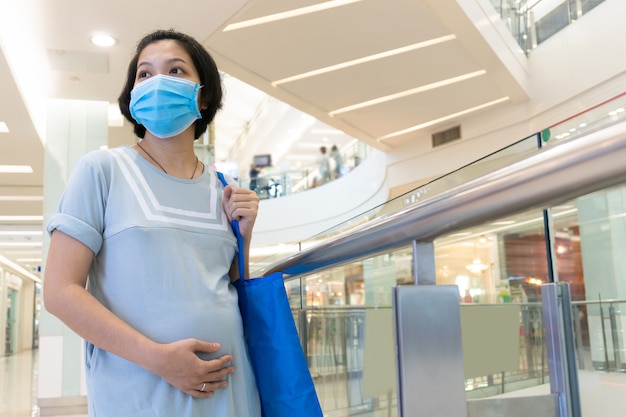 Mujer asiática embarazada con mascarilla y tocar el vientre en el centro comercial. Nuevo concepto de vida normal.