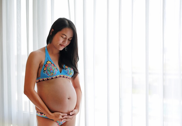 Mujer asiática embarazada que se coloca cerca de ventana en casa con mirar su vientre.