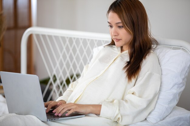 Mujer asiática embarazada usando la computadora portátil para trabajar en la cama en casa