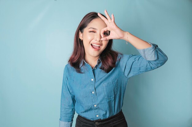 Mujer asiática emocionada con una camisa azul dando un gesto de mano OK aislado por un fondo azul