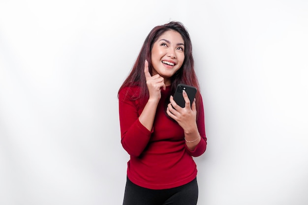Una mujer asiática emocionada con una camiseta roja apuntando al espacio de la copia encima de ella mientras sostenía su teléfono aislado por un fondo blanco