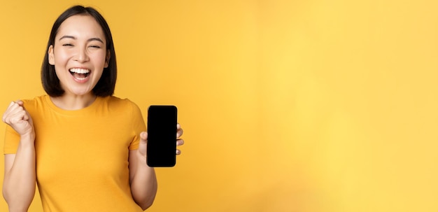 Mujer asiática emocionada mostrando la aplicación de teléfono inteligente y celebrando triunfante en el teléfono móvil de pie