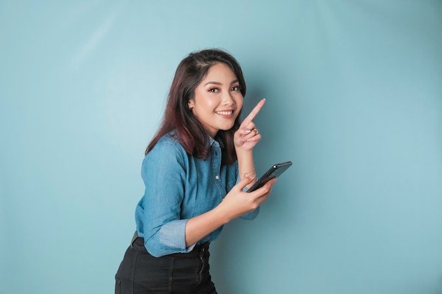 Mujer asiática emocionada con pantalones azul apuntando al espacio de copia a su lado mientras sostiene un teléfono inteligente aislado por fondo azul