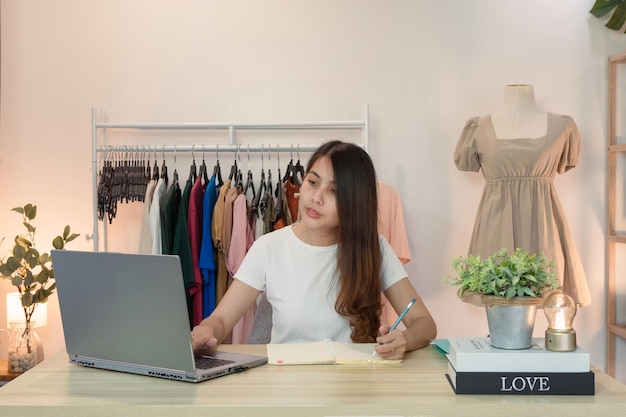 Mujer asiática emprendedora de tienda de ropa en línea usando laptop y tomando nota en el cuaderno en el escritorio en la oficina en casa