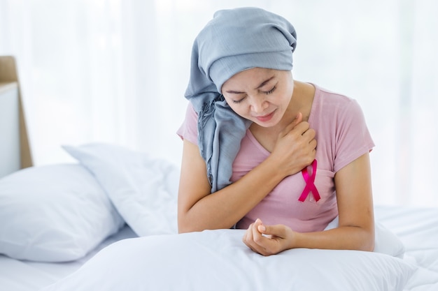 Una mujer asiática enfermedad paciente con cáncer de mama síntoma de ataque cardíaco agudo palo cinta rosa con pañuelo en la cabeza después del tratamiento de quimioterapia sentarse en la cama en el dormitorio de la casa