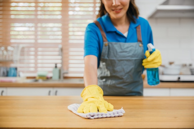 Mujer asiática enfocada con guantes amarillos limpia el mostrador de la cocina usando spray líquido y trapo Servicio de limpieza que garantiza la limpieza en el hogar Limpieza y desinfección del trabajo doméstico de la empleada doméstica