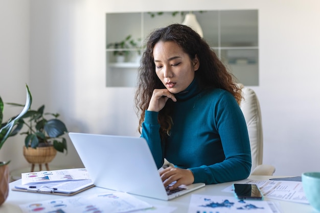 Mujer asiática enfocada usando una laptop en casa mirando la pantalla charlando leyendo o escribiendo correos electrónicos sentada en el sofá estudiante haciendo deberes trabajando en un proyecto de investigación en línea