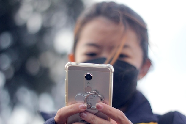 una mujer asiática enmascarada sostiene un teléfono móvil o un teléfono inteligente en la mano