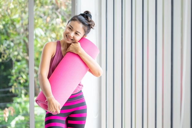 Una mujer asiática entrena practicando yoga con vestido rosa. Abraza la colchoneta de yoga rosa y practica la meditación, el estilo de vida de bienestar y el concepto de salud y fitness en un gimnasio.