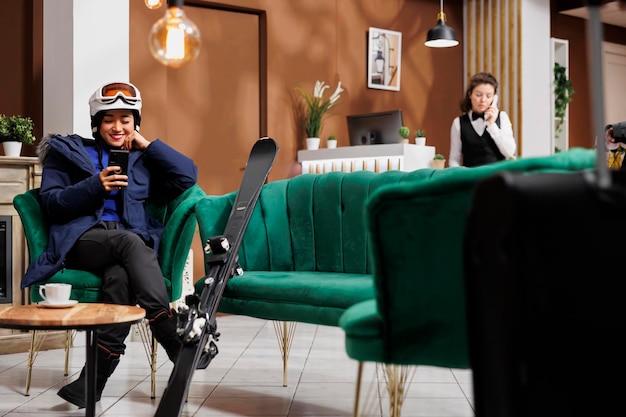 Mujer asiática entusiasta usando su teléfono celular y esperando en el vestíbulo del hotel de esquí para el procedimiento de reserva para las vacaciones de invierno Viajero en equipo de deportes de invierno con su teléfono inteligente en el área de salón de la estación de esquí