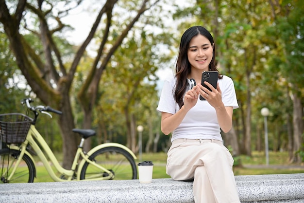 Una mujer asiática escalofriante se relaja sentada en un banco y usando su teléfono inteligente en el parque verde