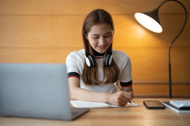 Mujer asiática escribiendo haciendo una lista tomando notas en el bloc de notas trabajando o aprendiendo en una computadora portátil en interiores curso educativo o seminario de capacitación educación concepto en línea