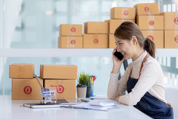 Una mujer asiática está hablando por teléfono con un cliente para confirmar un pedido. Posee una tienda en línea que empaca y envía a través de una empresa de transporte privada Conceptos de venta y compras en línea