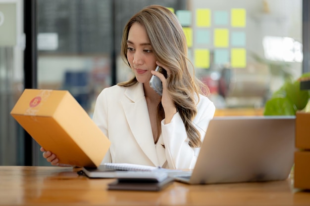 Una mujer asiática está hablando por teléfono con un cliente para confirmar un pedido. Posee una tienda en línea que empaca y envía a través de una empresa de transporte privada Conceptos de venta y compras en línea