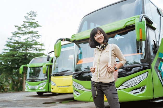 Una mujer asiática está de pie en una mochila y auriculares sonriendo con el telón de fondo del autobús