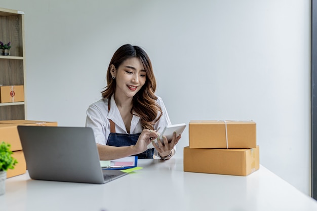 Una mujer asiática está presionando una calculadora blanca, es dueña de una tienda en línea, está usando una calculadora de monto de pedido para obtener un resumen del monto que los clientes deben pagar. Concepto de venta online.