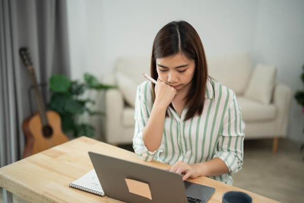 Una mujer asiática está sentada en un escritorio con una computadora portátil y un cuaderno está mirando la pantalla de la computadora portable y está profundamente pensativa