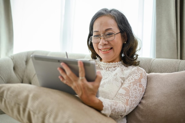 Una mujer asiática feliz con anteojos se sienta en un sofá en la sala de estar usando una tableta digital