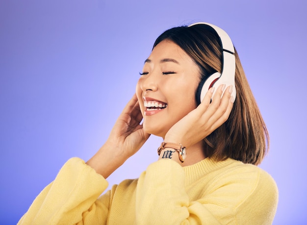 Mujer asiática feliz con auriculares y escuchando música para transmisión en línea contra un fondo de estudio púrpura Persona femenina o modelo sonriendo disfrutando del sonido de la pista de audio o canciones en auriculares en maqueta