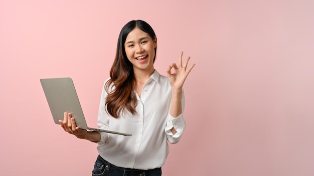 Mujer asiática feliz en una camisa blanca con su computadora portátil que muestra el signo de la mano bien