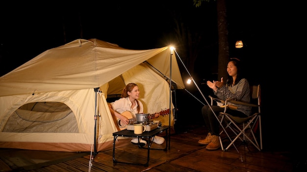 Una mujer asiática feliz canta una canción y toca la guitarra con sus amigos en el campamento por la noche