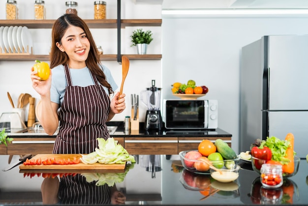 Mujer asiática feliz con delantal sosteniendo ají y espátula de madera Preparando una ensalada saludable con verduras frescas como tomate de zanahoria y roble verde en la cocina casera