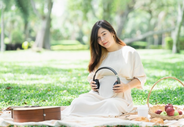 Mujer asiática feliz embarazada poniendo auriculares en su vientre para escuchar música mientras se sienta en una manta de picnic en el césped en el parque del jardín bajo el árbol Familia madre mamá embarazada concepto saludable