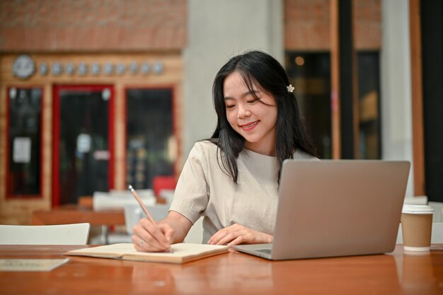 Una mujer asiática feliz está escribiendo algo en su libro mientras trabaja de forma remota en una cafetería