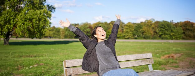 Foto mujer asiática feliz estirando sus manos sentada en un banco con rostro emocionado sonriendo sensación de satisfacción