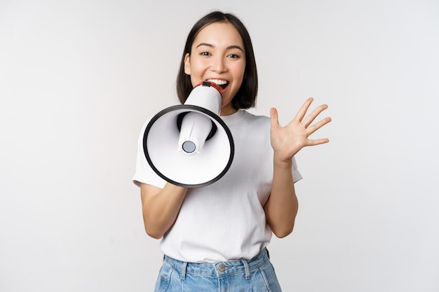 Mujer asiática feliz gritando al megáfono haciendo un anuncio anunciando algo de pie sobre fondo blanco.