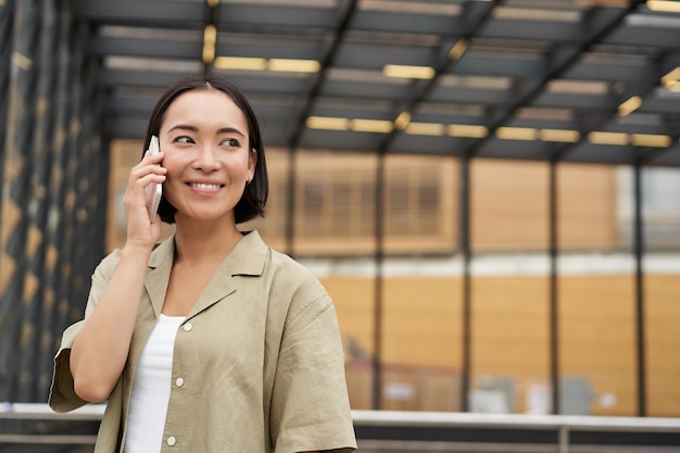 Mujer asiática feliz hablando por teléfono móvil de pie en la parada de autobús en la ciudad al aire libre